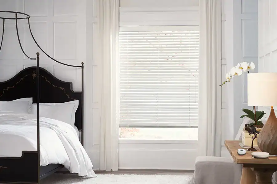 Bright, modern bedroom with a large window covered by white blinds and sheer curtains. Featuring a black canopy bed with white bedding, a wooden desk, and elegant floors crafted by a top flooring company Suffolk County, NY.