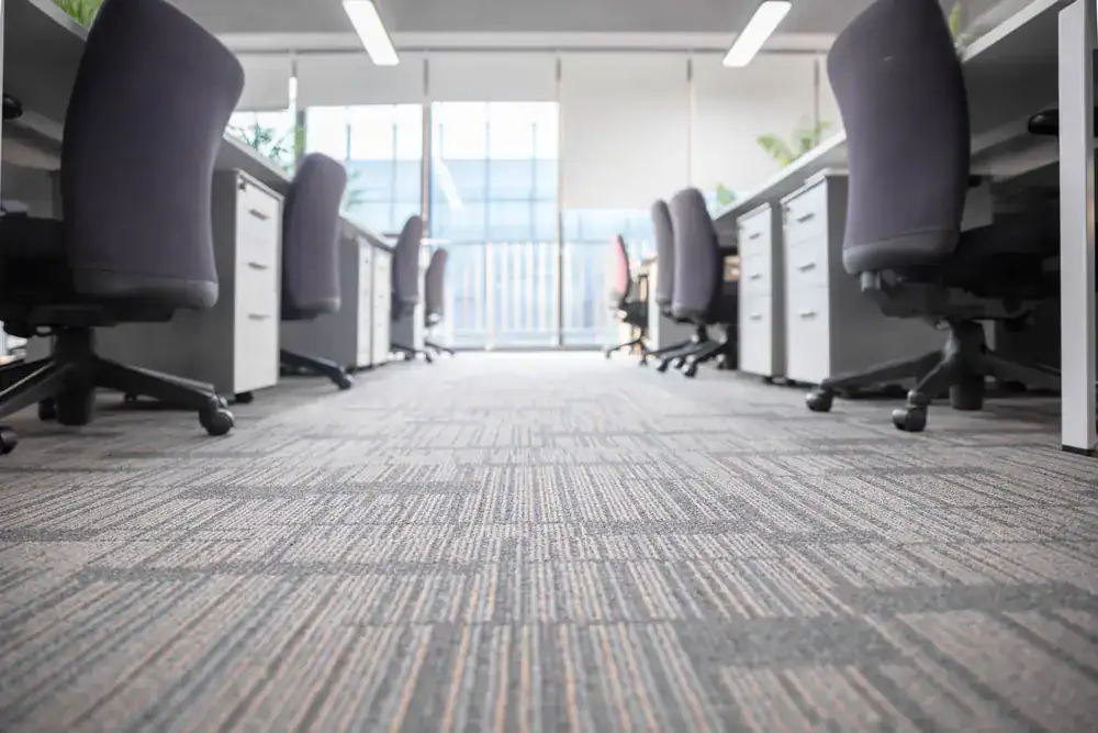A row of empty office chairs and desks lines both sides of a carpeted hallway in a bright, modern office with large windows—the work of a top flooring company in Suffolk County, NY.