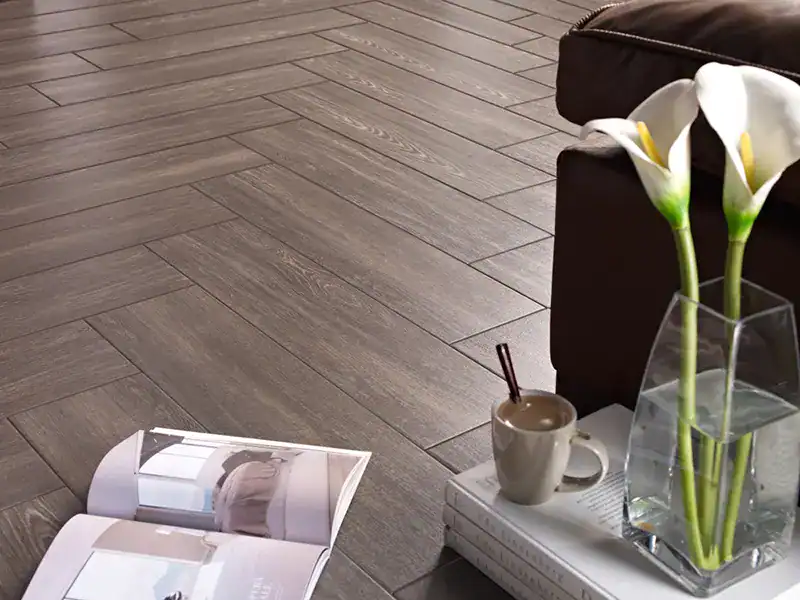 A cozy scene with a brown herringbone wood floor from a top flooring company in Suffolk County, NY, an open magazine, a cup of coffee on two stacked books, and a vase with white calla lilies next to a brown sofa.